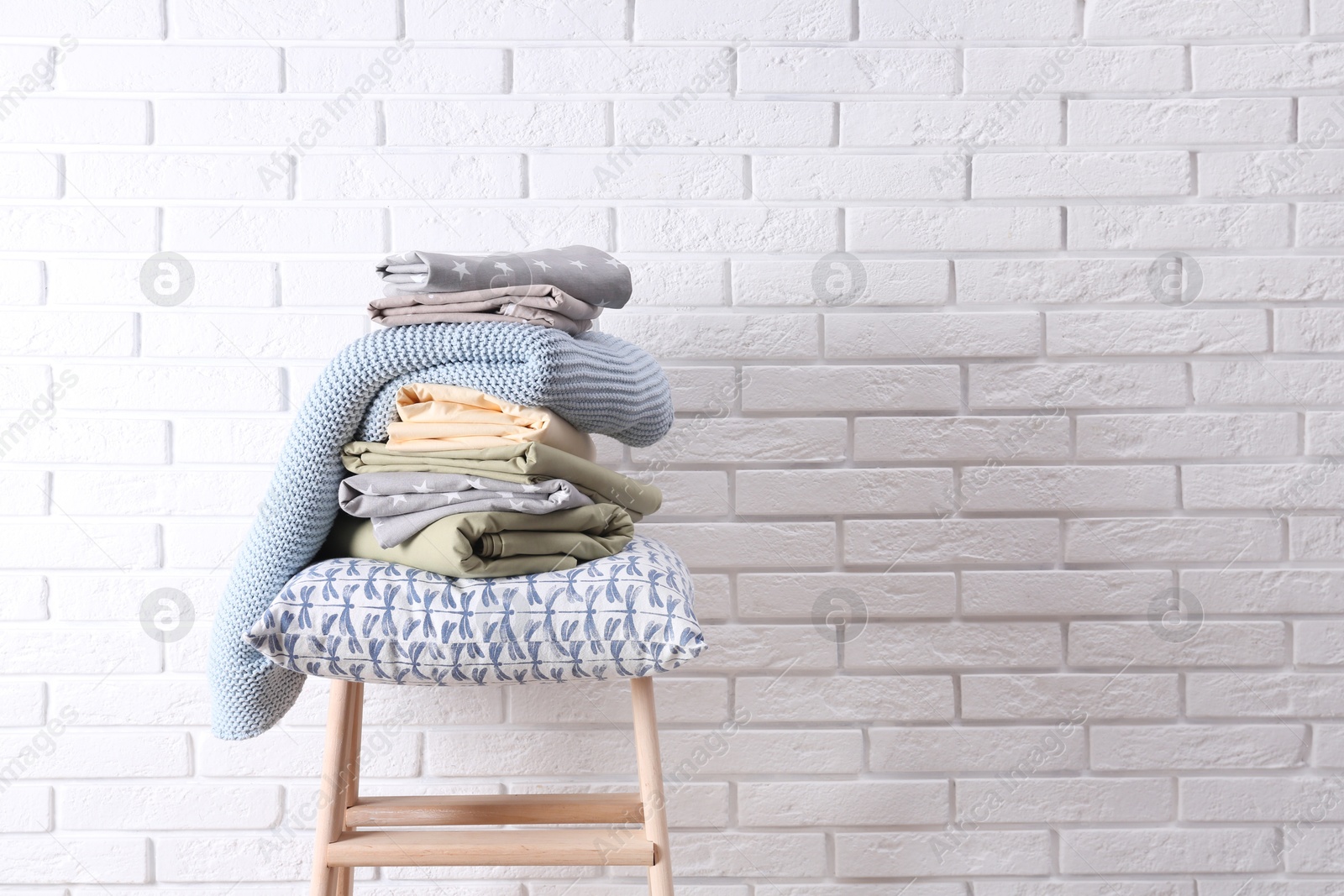 Photo of Stack of clean bed sheets and pillow on stool near white brick wall. Space for text