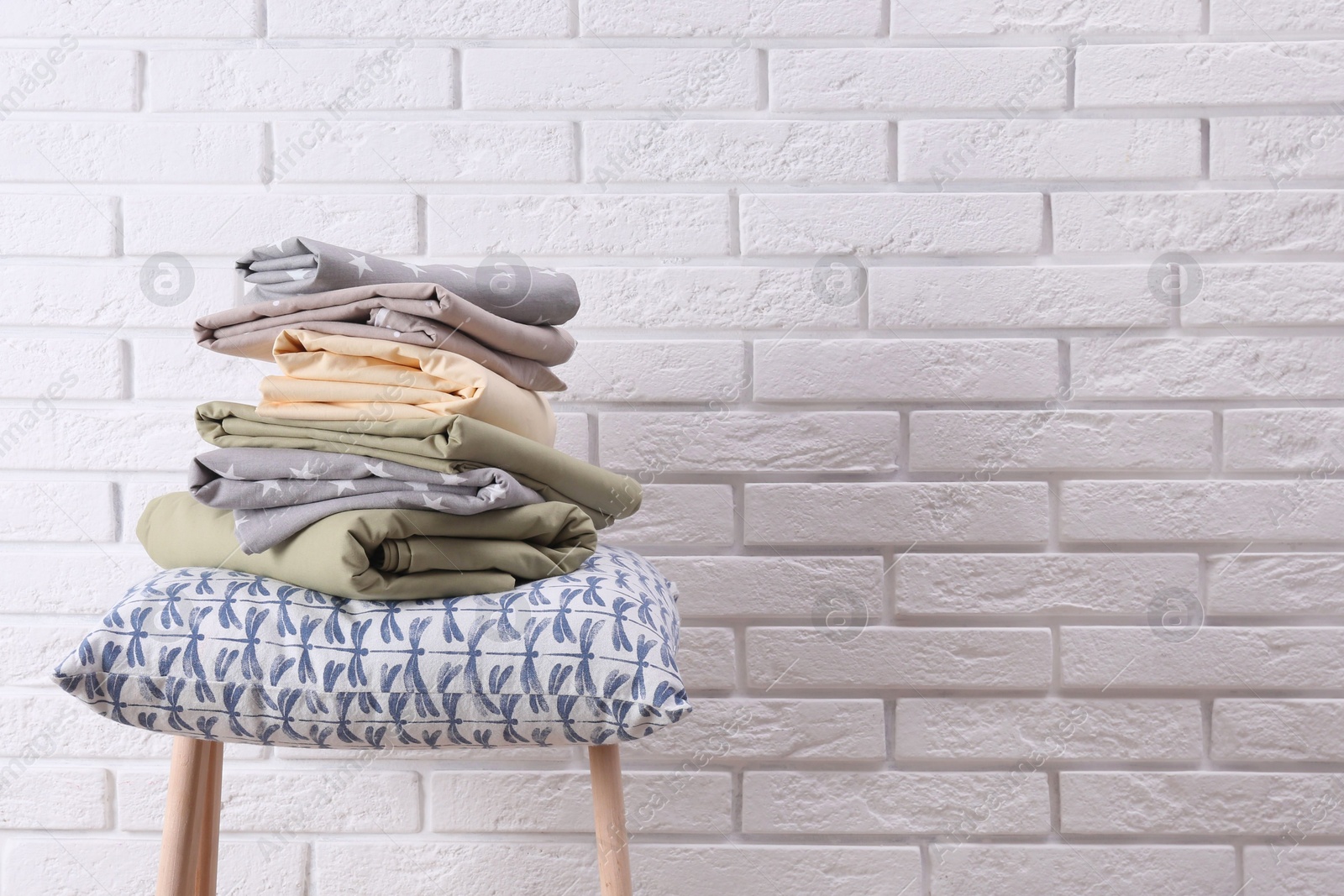 Photo of Stack of clean bed sheets and pillow on stool near white brick wall. Space for text