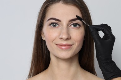 Photo of Beautician brushing client's eyebrows after henna dyeing procedure on light background, closeup