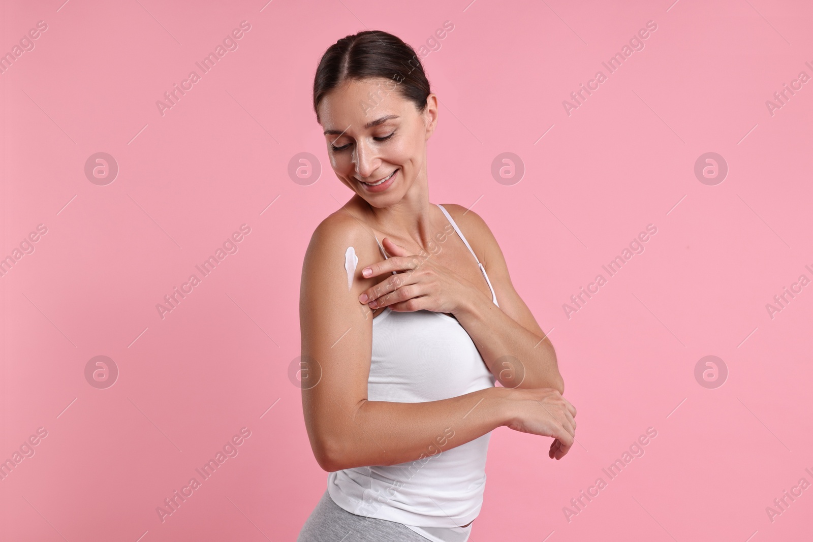 Photo of Smiling woman applying cream onto shoulder against pink background. Body care