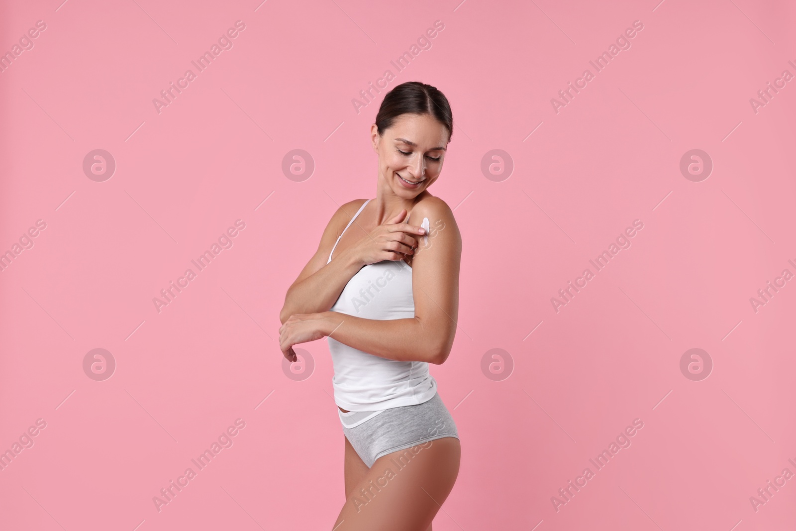 Photo of Smiling woman applying cream onto shoulder against pink background. Body care