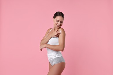 Smiling woman applying cream onto shoulder against pink background. Body care