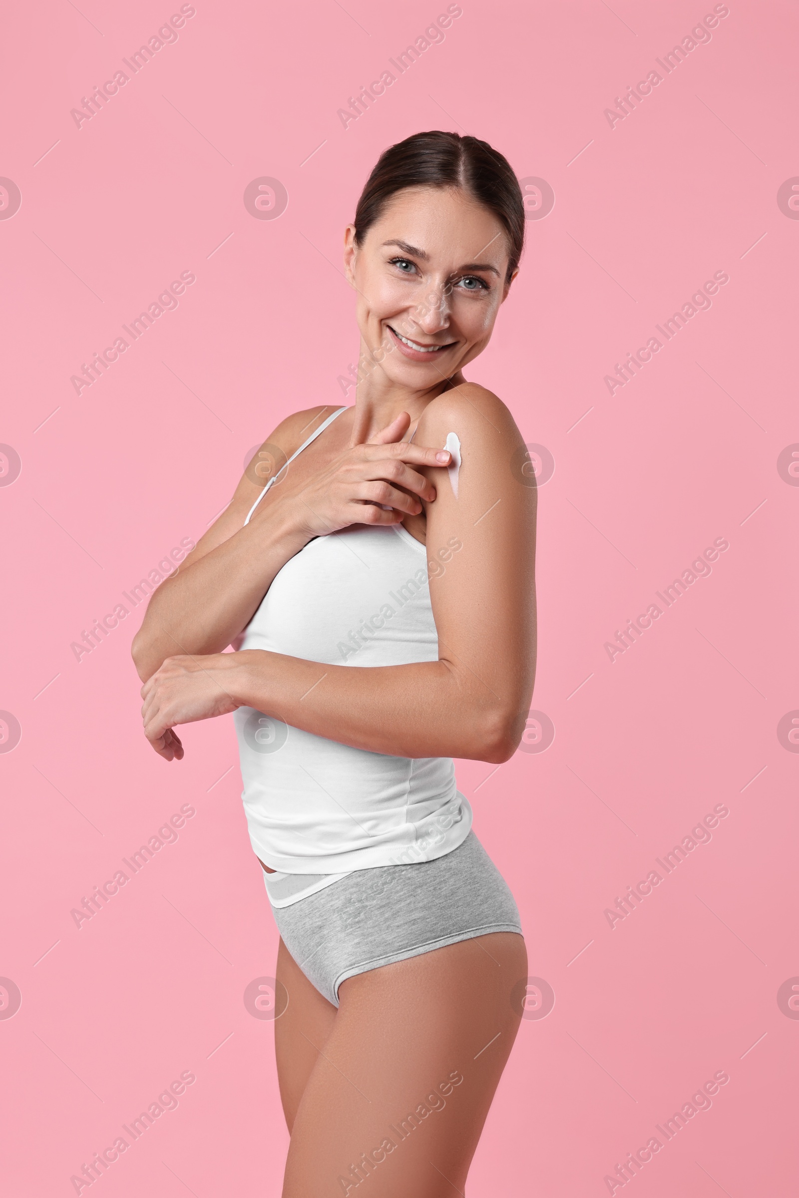 Photo of Smiling woman applying cream onto shoulder against pink background. Body care