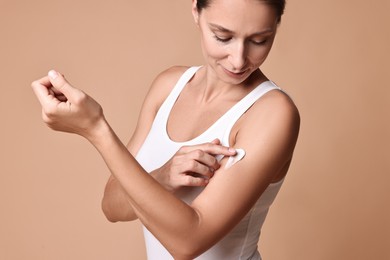 Beautiful woman applying cream onto arm against beige background. Body care