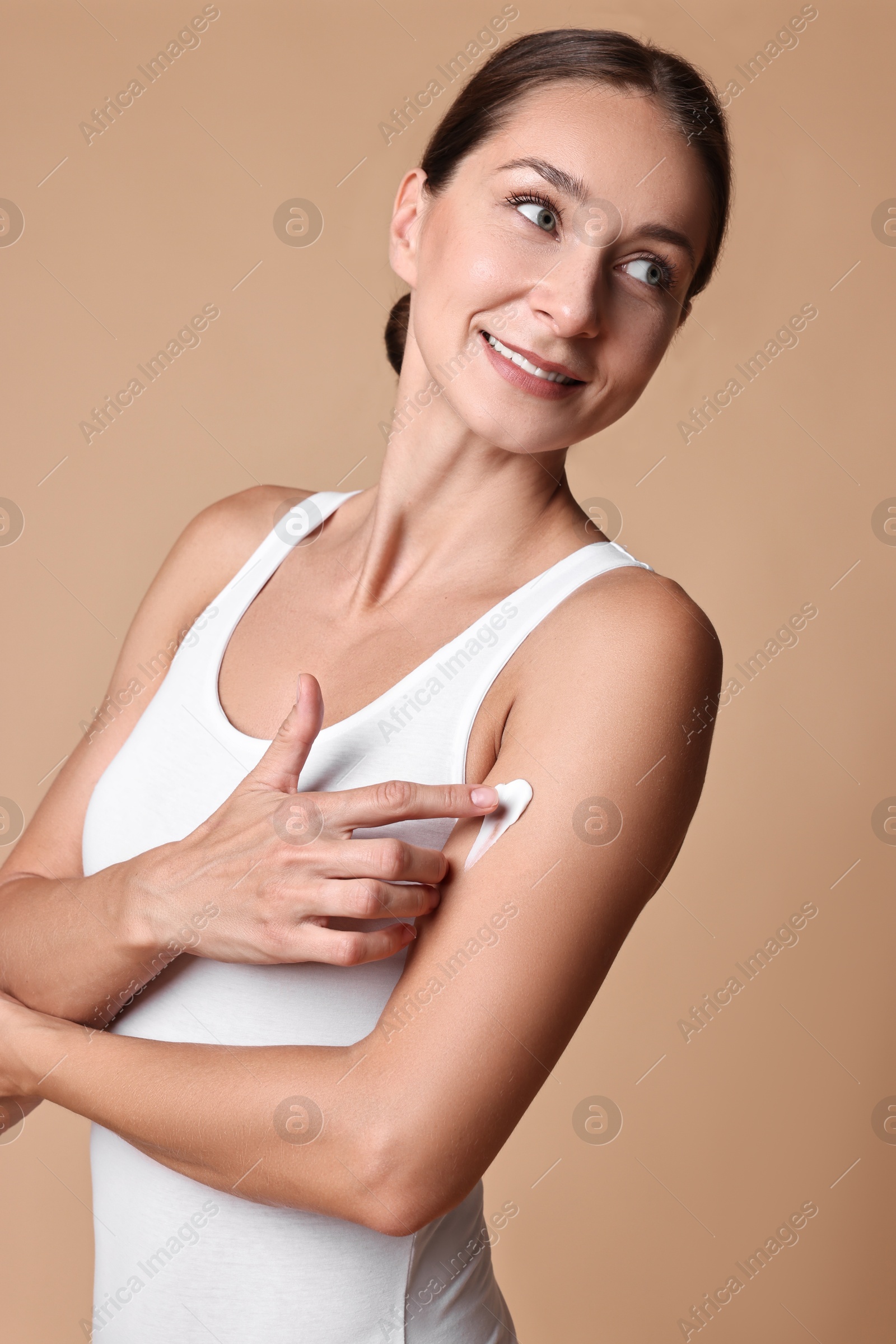 Photo of Smiling woman applying cream onto arm against beige background. Body care