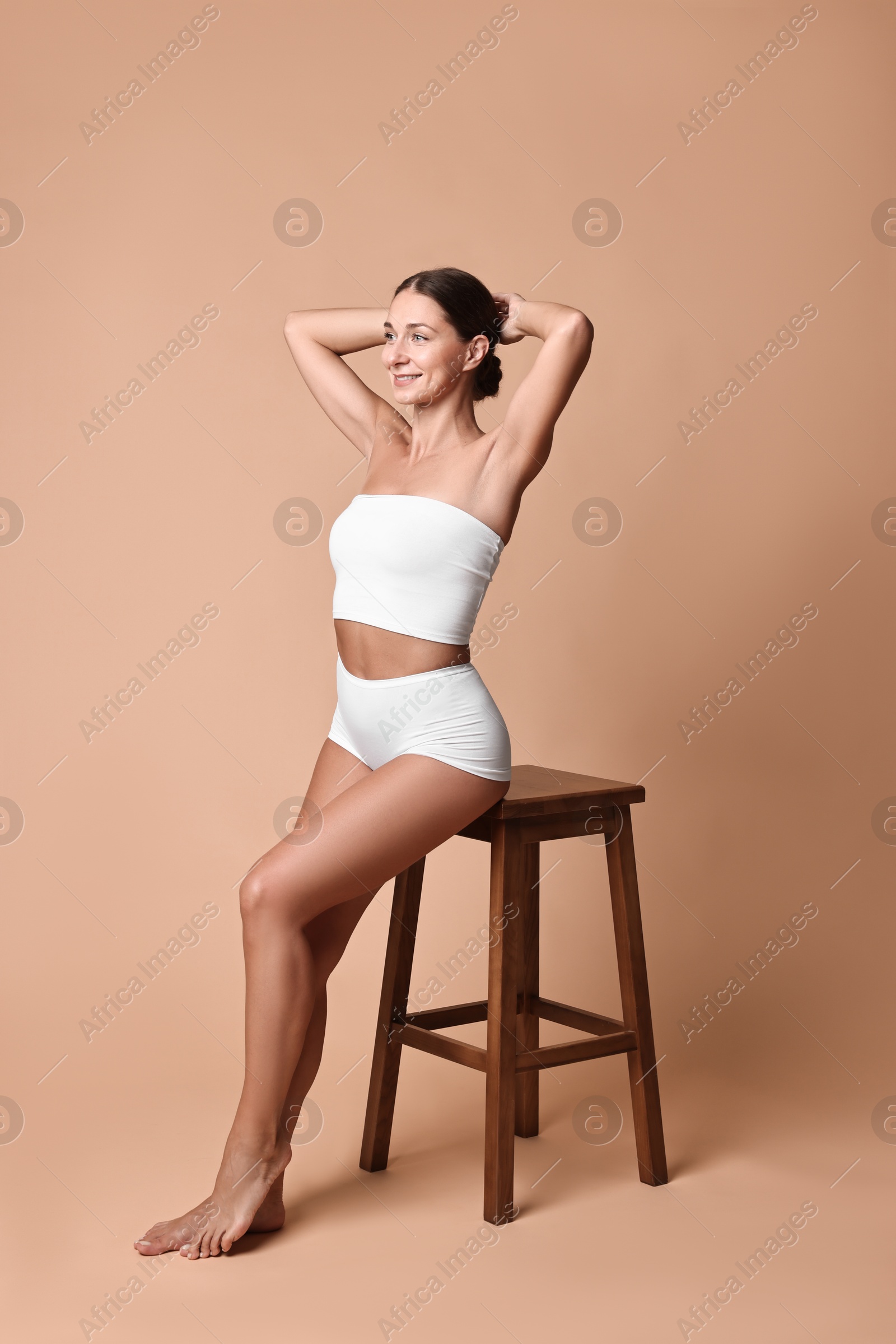 Photo of Smiling woman with perfect skin in underwear posing on stool against beige background. Body care