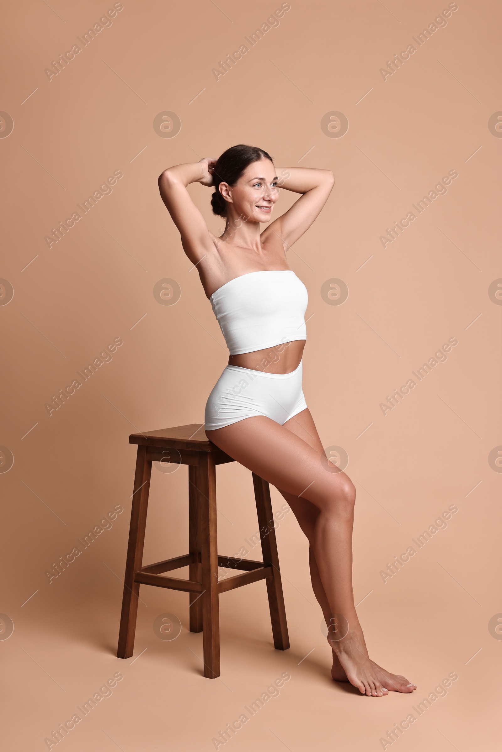 Photo of Smiling woman with perfect skin in underwear posing on stool against beige background. Body care