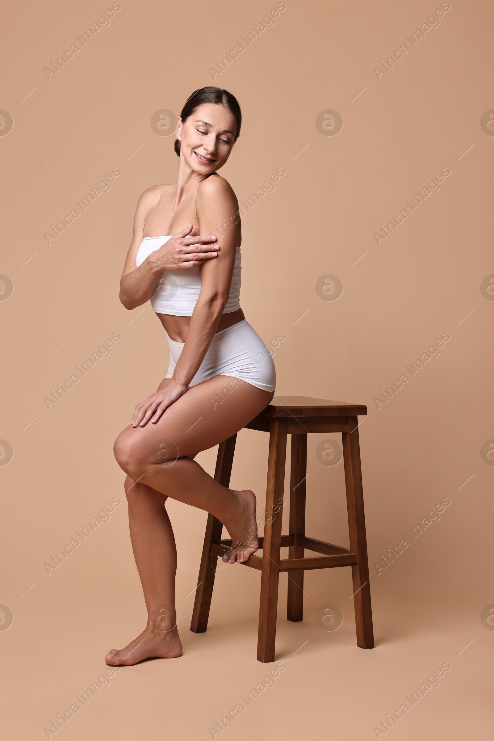 Photo of Smiling woman with perfect skin in underwear posing with stool against beige background. Body care