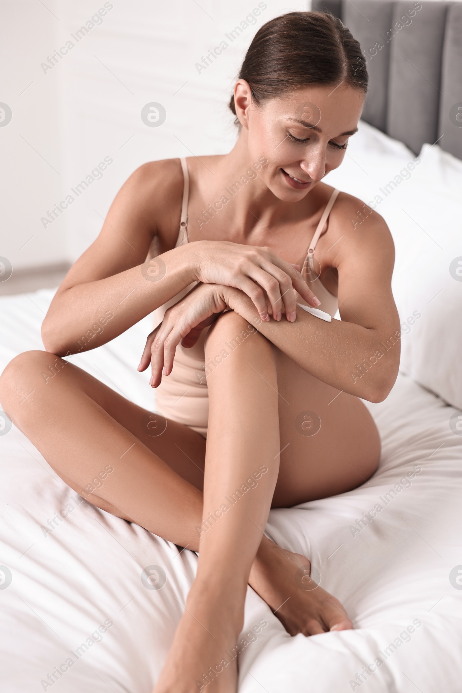 Photo of Smiling woman with perfect skin applying cream onto hand on bed. Body care