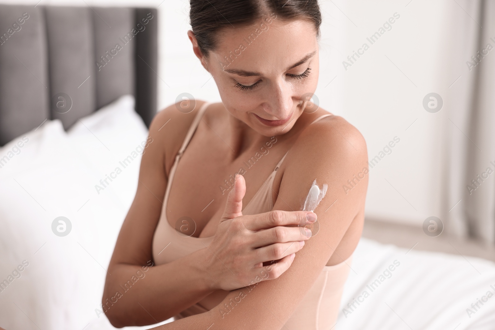 Photo of Beautiful woman with perfect skin applying cream onto shoulder on bed. Body care