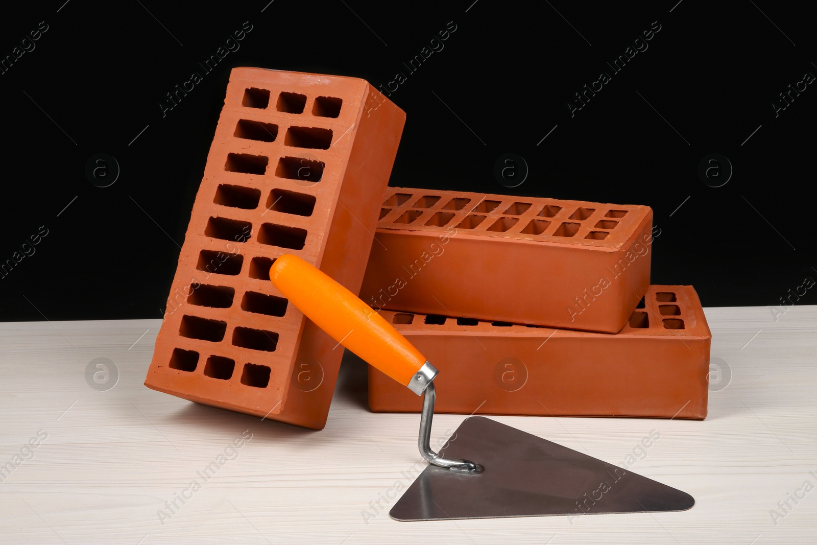 Photo of Many red bricks and putty knife on light wooden table against black background. Building material