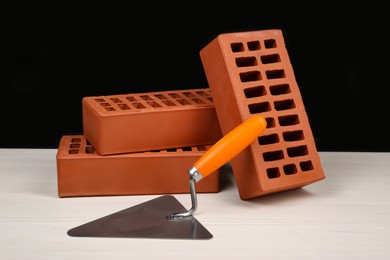 Photo of Many red bricks and putty knife on light wooden table against black background. Building material