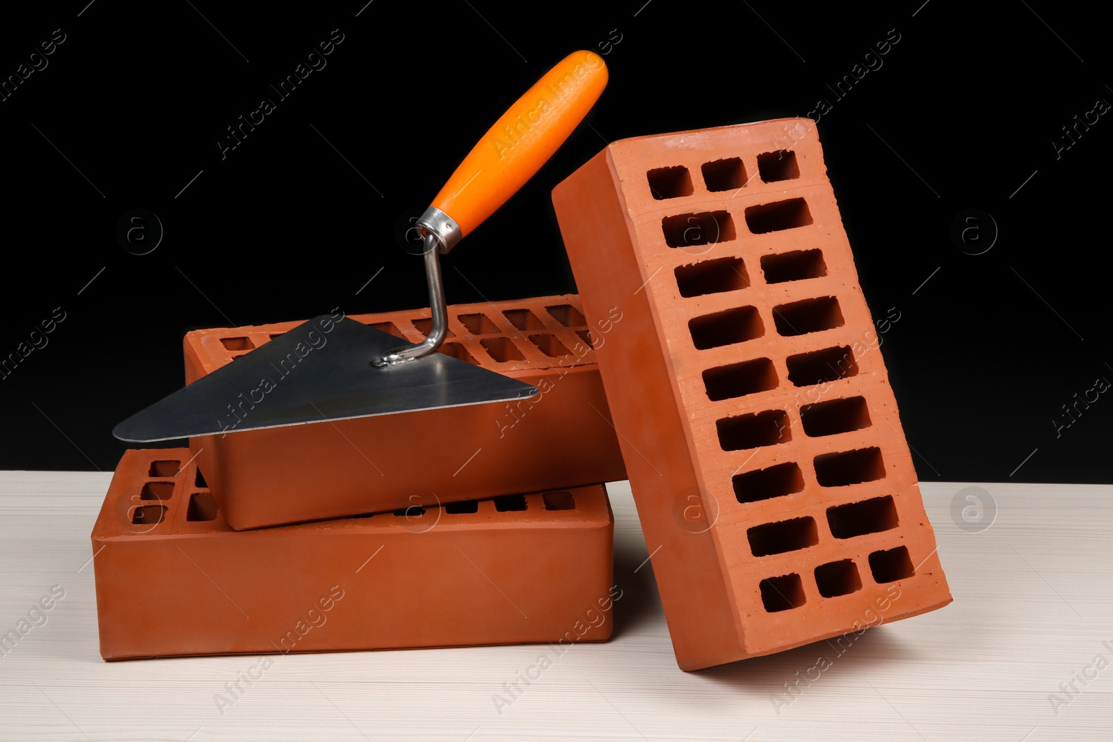 Photo of Many red bricks and putty knife on light wooden table against black background. Building material