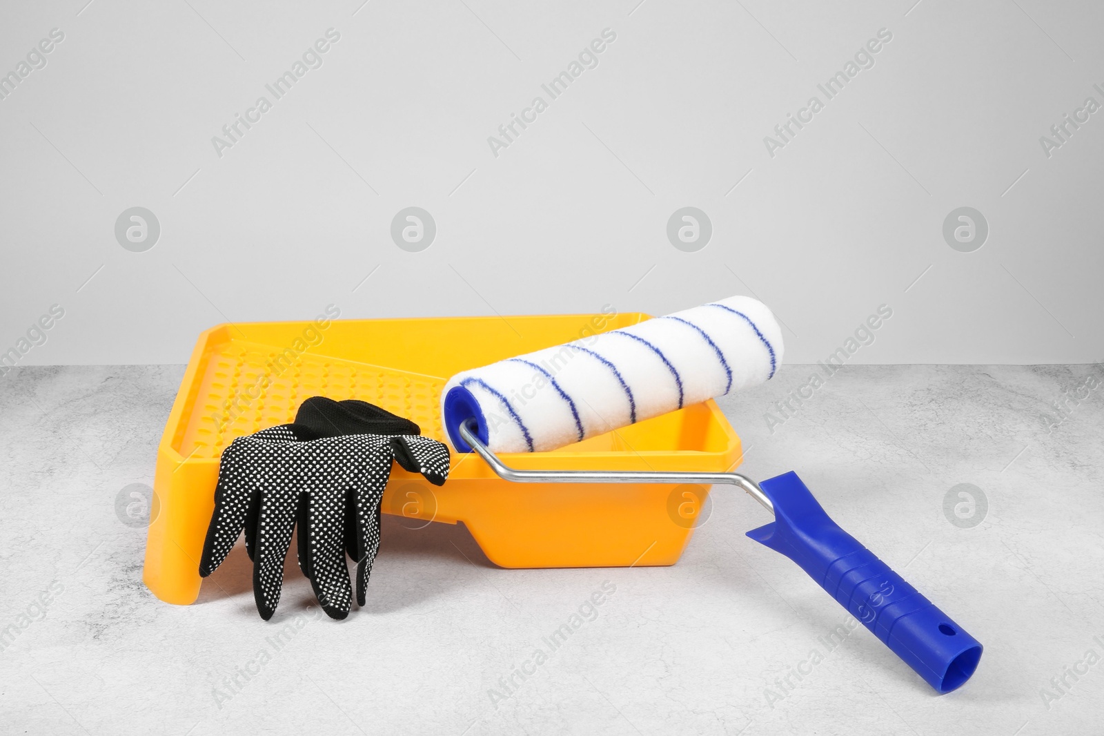 Photo of Paint roller, gloves and yellow tray on textured table against light background. Building material