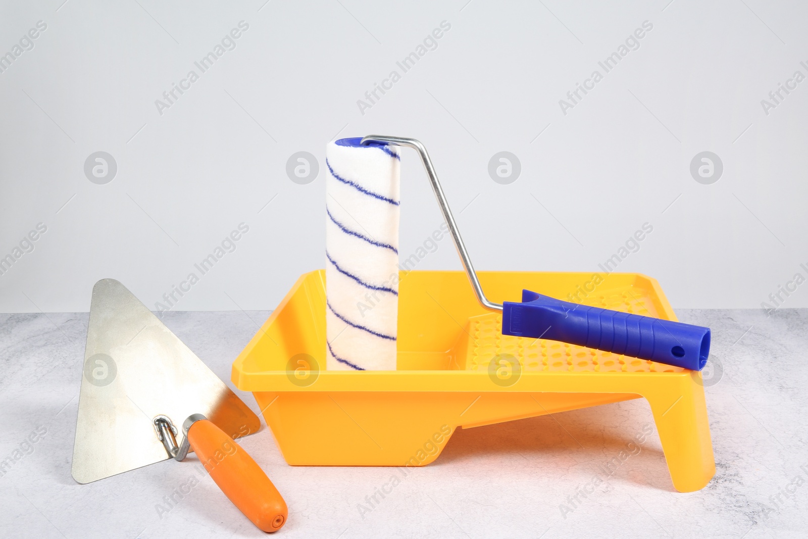 Photo of Paint roller, trowel and yellow tray on textured table against light background. Building material