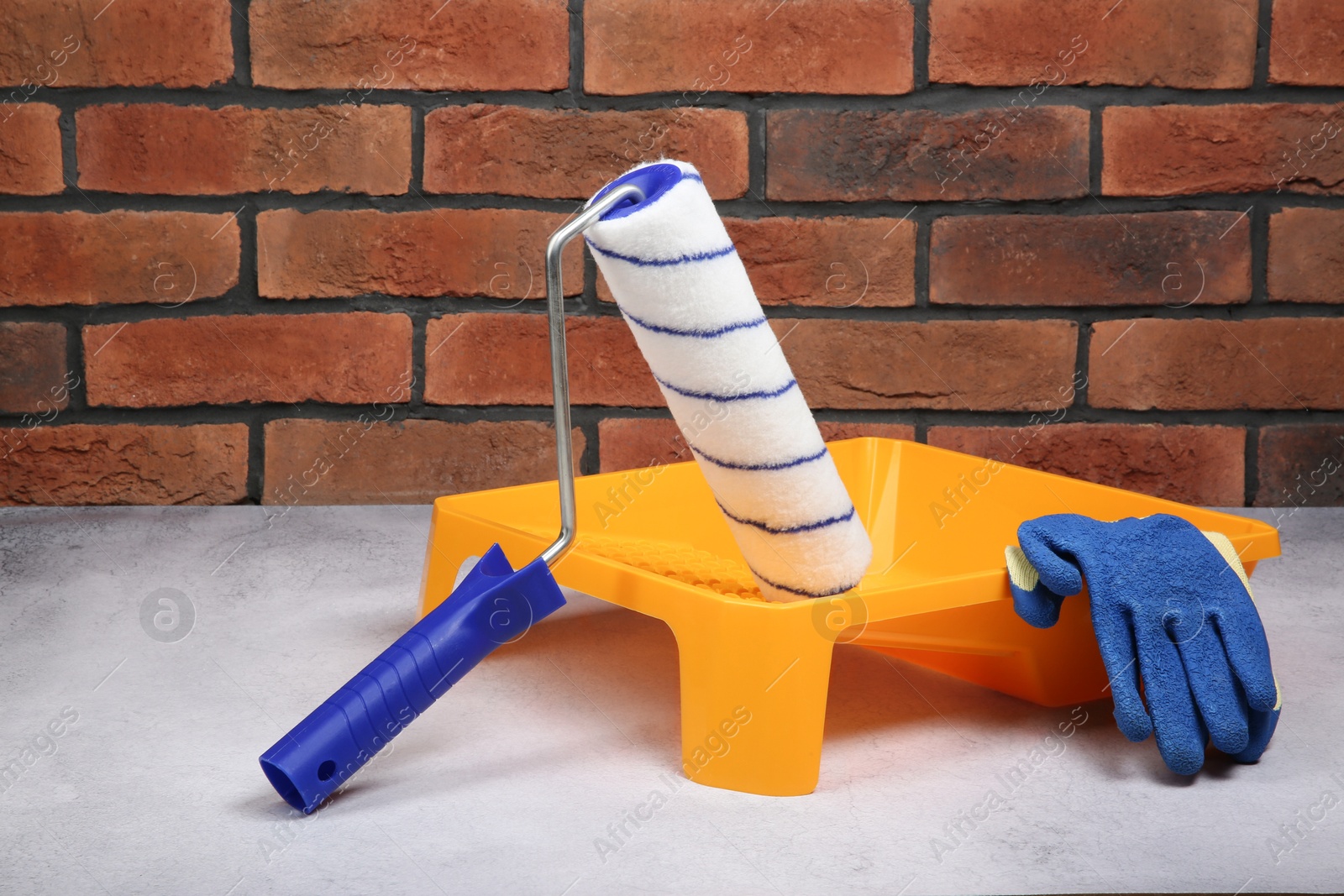 Photo of Paint roller, gloves and yellow tray on light textured table against red brick wall