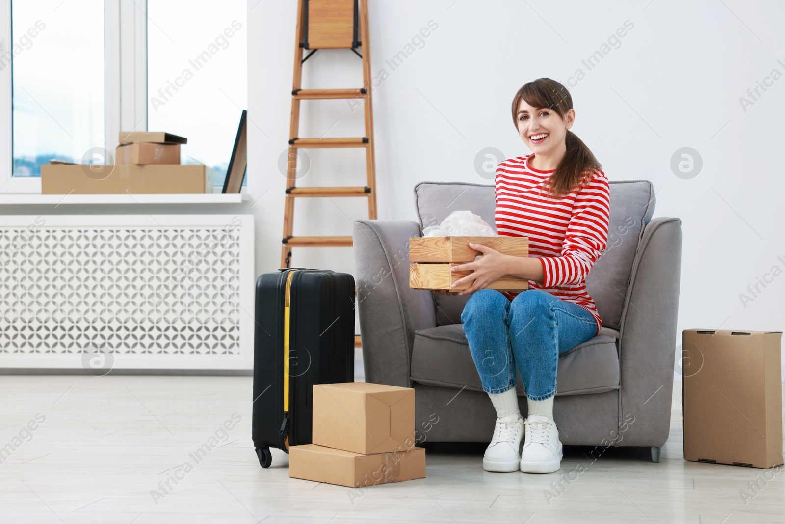 Photo of Happy woman with moving boxes and suitcase in new apartment. Housewarming party