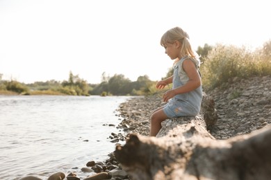 Photo of Cute little girl sitting on tree trunk near river. Child enjoying beautiful nature