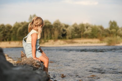 Photo of Cute little girl sitting on tree trunk near river. Child enjoying beautiful nature