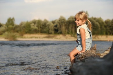 Photo of Cute little girl sitting on tree trunk near river. Child enjoying beautiful nature