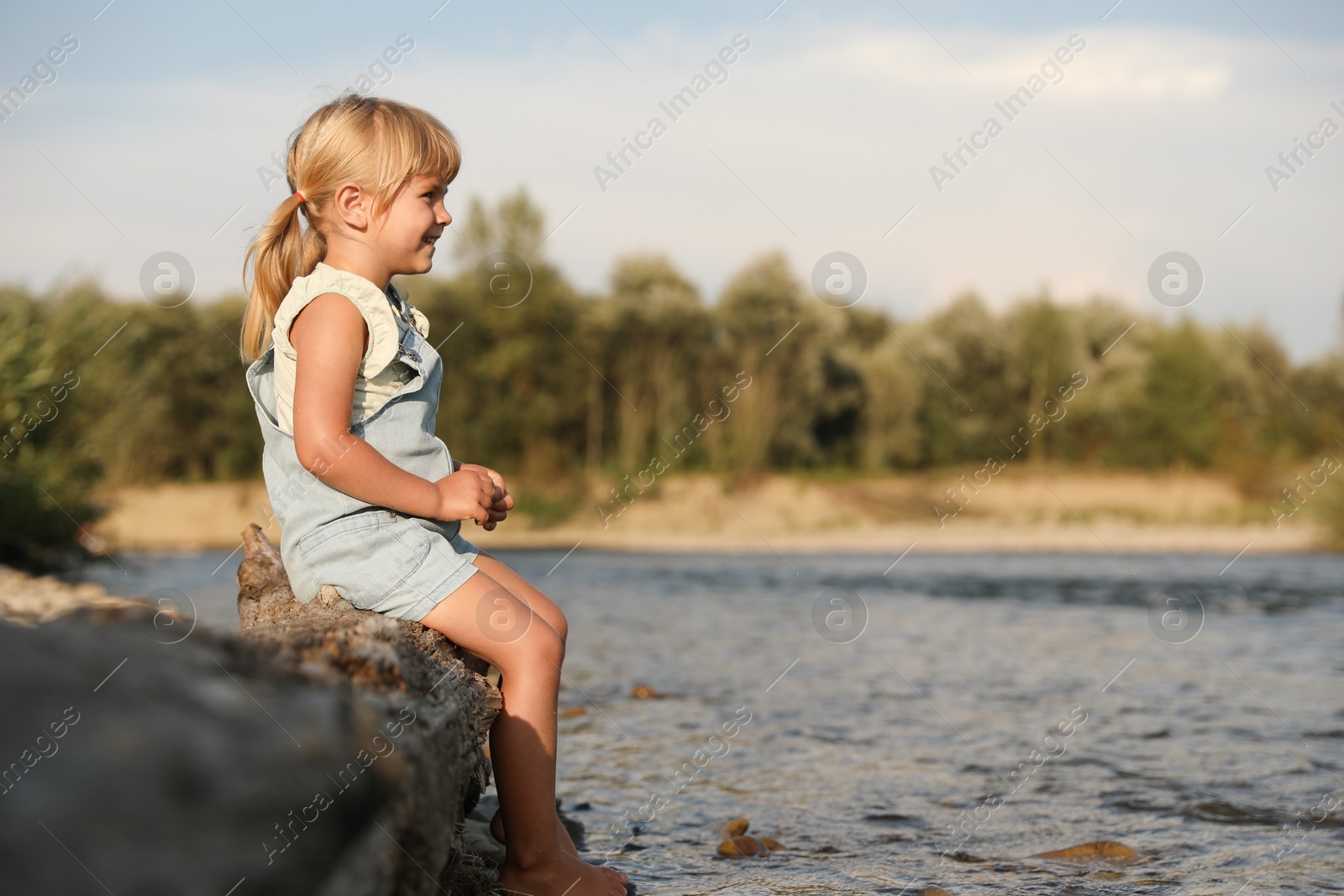 Photo of Cute little girl sitting on tree trunk near river. Child enjoying beautiful nature