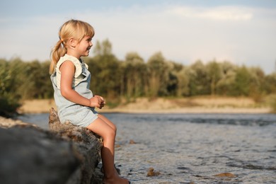 Cute little girl sitting on tree trunk near river. Child enjoying beautiful nature