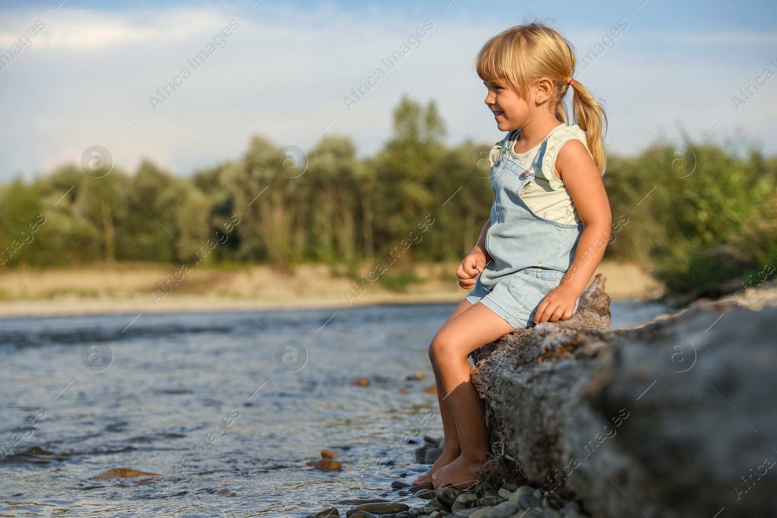 Photo of Cute little girl sitting on tree trunk near river. Child enjoying beautiful nature