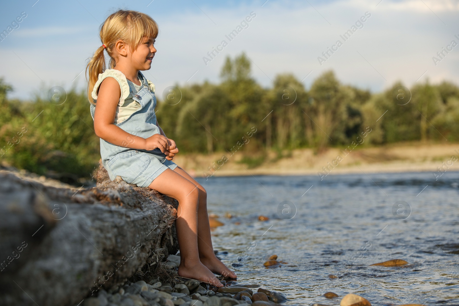 Photo of Cute little girl sitting on tree trunk near river. Child enjoying beautiful nature