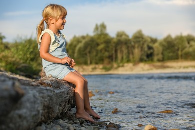 Photo of Cute little girl sitting on tree trunk near river. Child enjoying beautiful nature