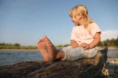 Cute little girl sitting on tree trunk near river, selective focus. Child enjoying beautiful nature