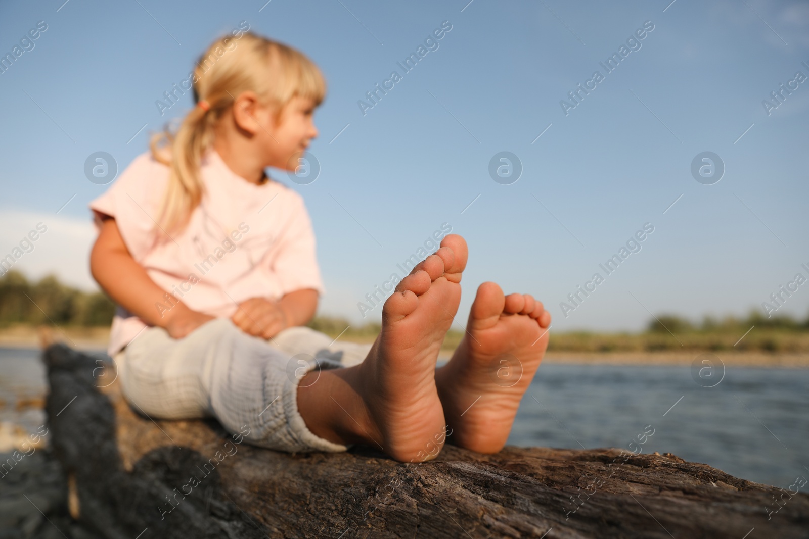 Photo of Cute little girl sitting on tree trunk near river, selective focus. Child enjoying beautiful nature