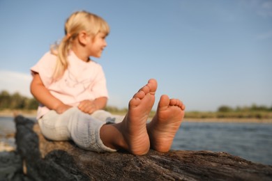 Photo of Cute little girl sitting on tree trunk near river, selective focus. Child enjoying beautiful nature