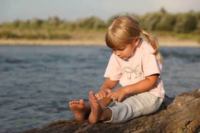 Photo of Cute little girl sitting on tree trunk near river, space for text. Child enjoying beautiful nature