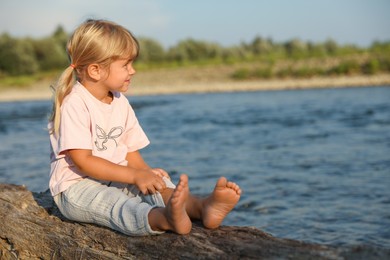 Photo of Cute little girl sitting on tree trunk near river, space for text. Child enjoying beautiful nature