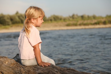 Photo of Cute little girl sitting on tree trunk near river, space for text. Child enjoying beautiful nature