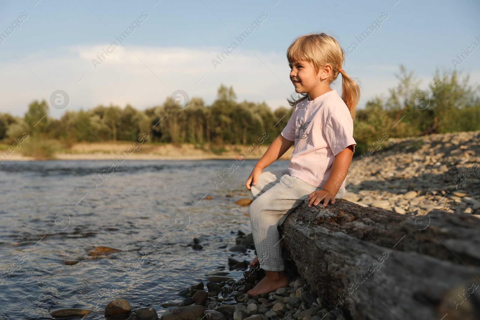 Photo of Cute little girl sitting on tree trunk near river. Child enjoying beautiful nature