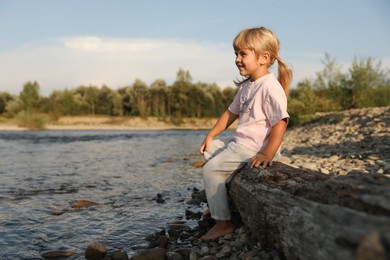 Cute little girl sitting on tree trunk near river. Child enjoying beautiful nature