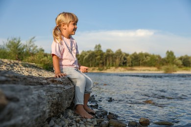 Photo of Cute little girl sitting on tree trunk near river. Child enjoying beautiful nature