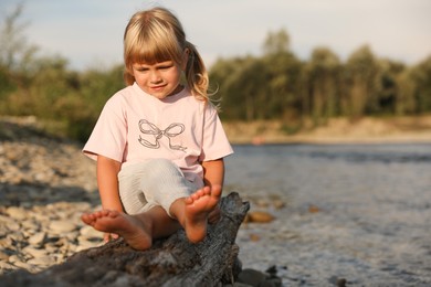 Photo of Cute little girl sitting on tree trunk near river. Child enjoying beautiful nature