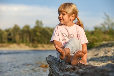 Photo of Cute little girl sitting on tree trunk near river. Child enjoying beautiful nature