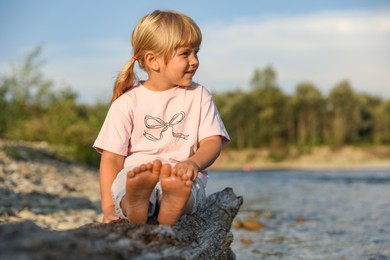 Photo of Cute little girl sitting on tree trunk near river. Child enjoying beautiful nature