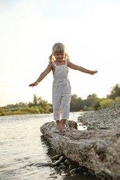 Photo of Cute little girl walking along tree trunk near river. Child enjoying beautiful nature