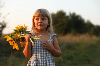 Photo of Little girl with sunflowers at meadow, space for text. Child enjoying beautiful nature