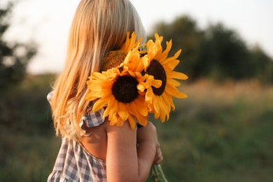 Photo of Little girl with sunflowers at meadow, space for text. Child enjoying beautiful nature