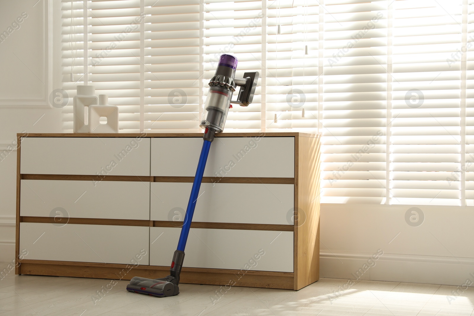 Photo of One cordless vacuum cleaner leaning on chest of drawers indoors