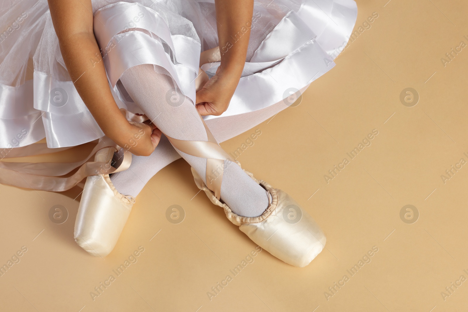 Photo of Little ballerina tying pointe shoes on beige background, closeup