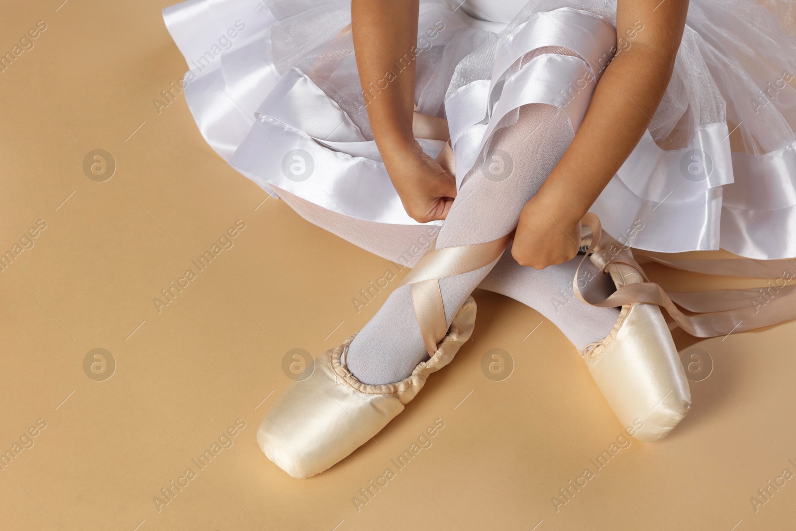 Photo of Little ballerina tying pointe shoes on beige background, closeup