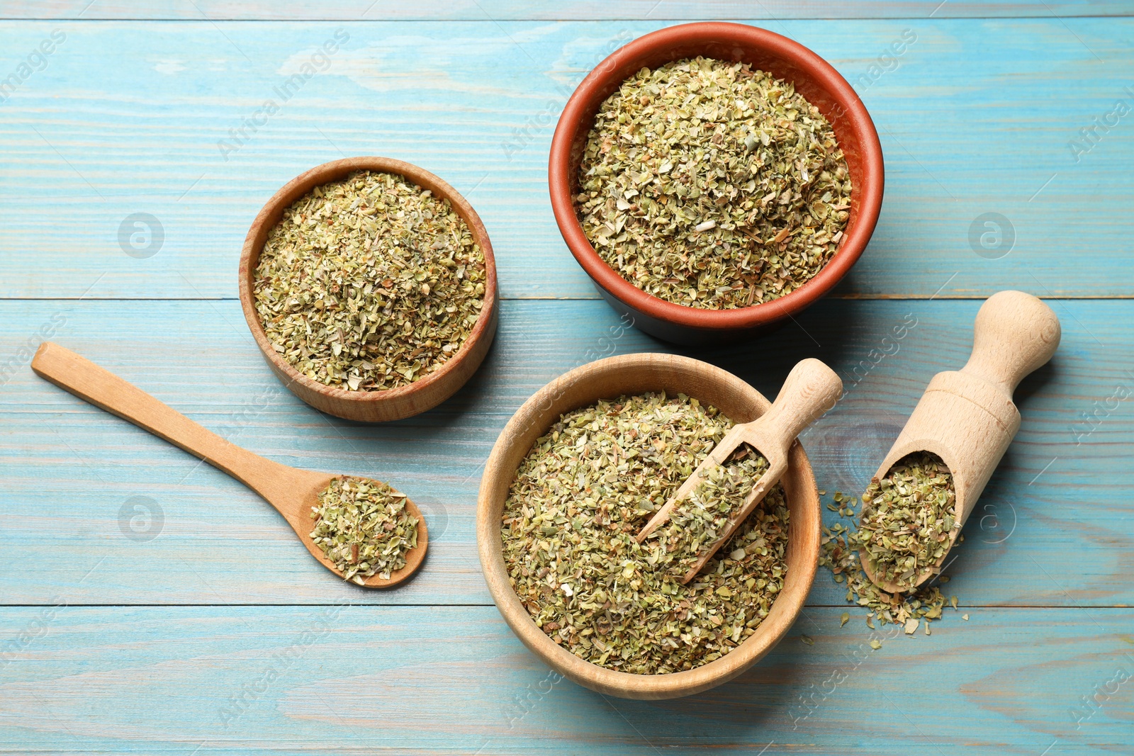 Photo of Dried oregano in bowls, scoops and spoon on light blue wooden table, flat lay