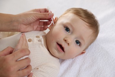 Mother cleaning ear of her cute little baby with cotton swab on bed, closeup