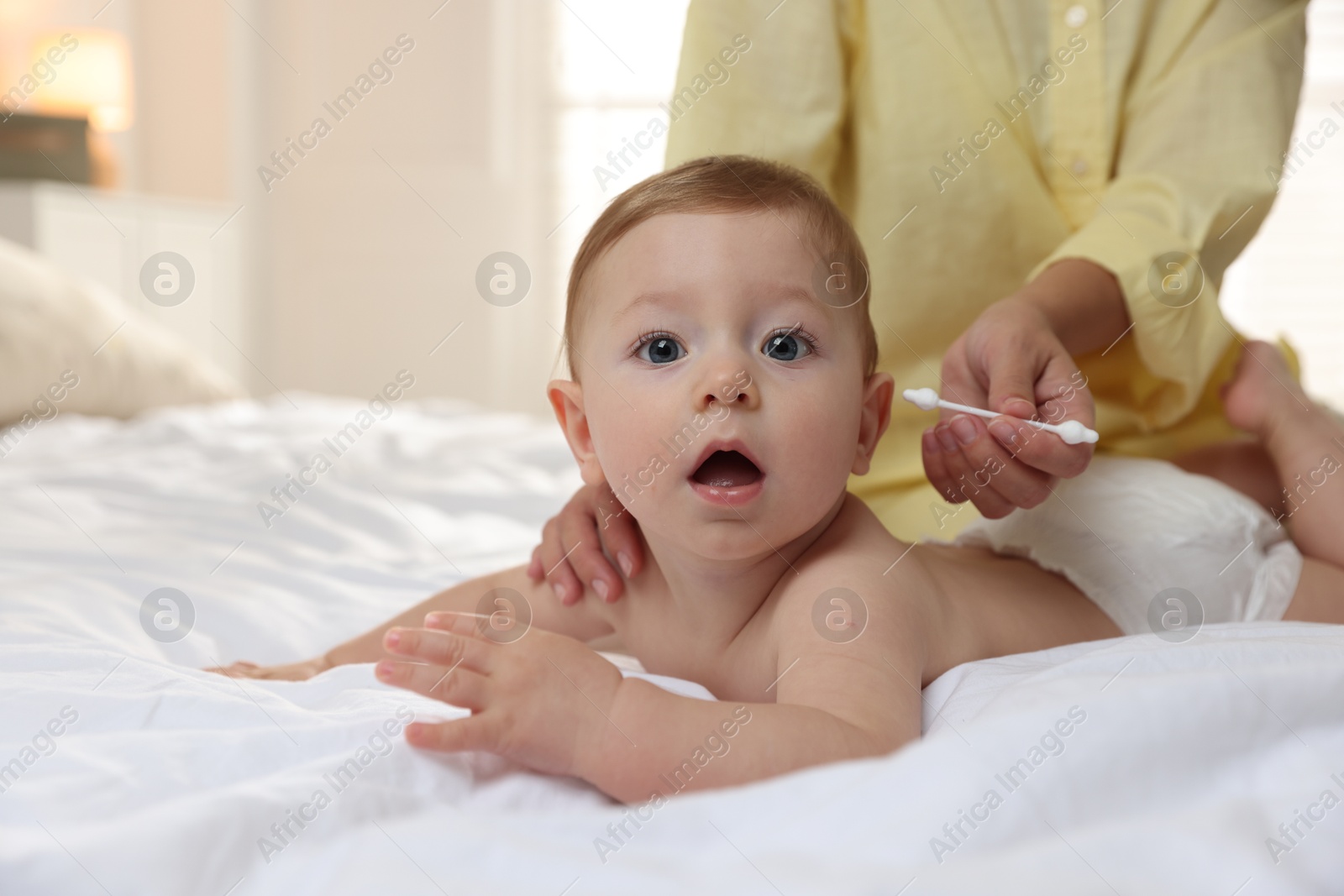 Photo of Mother cleaning ear of her cute little baby with cotton swab on bed, closeup